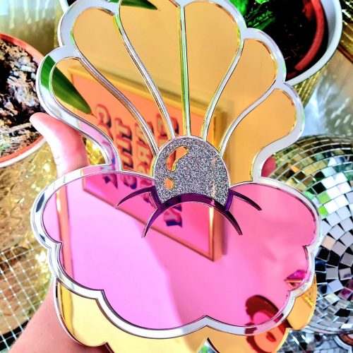 A handmade mirror in the shapeof a clam shell. The clam has a silver outline, gold petals and a glitter pearl in the centre.