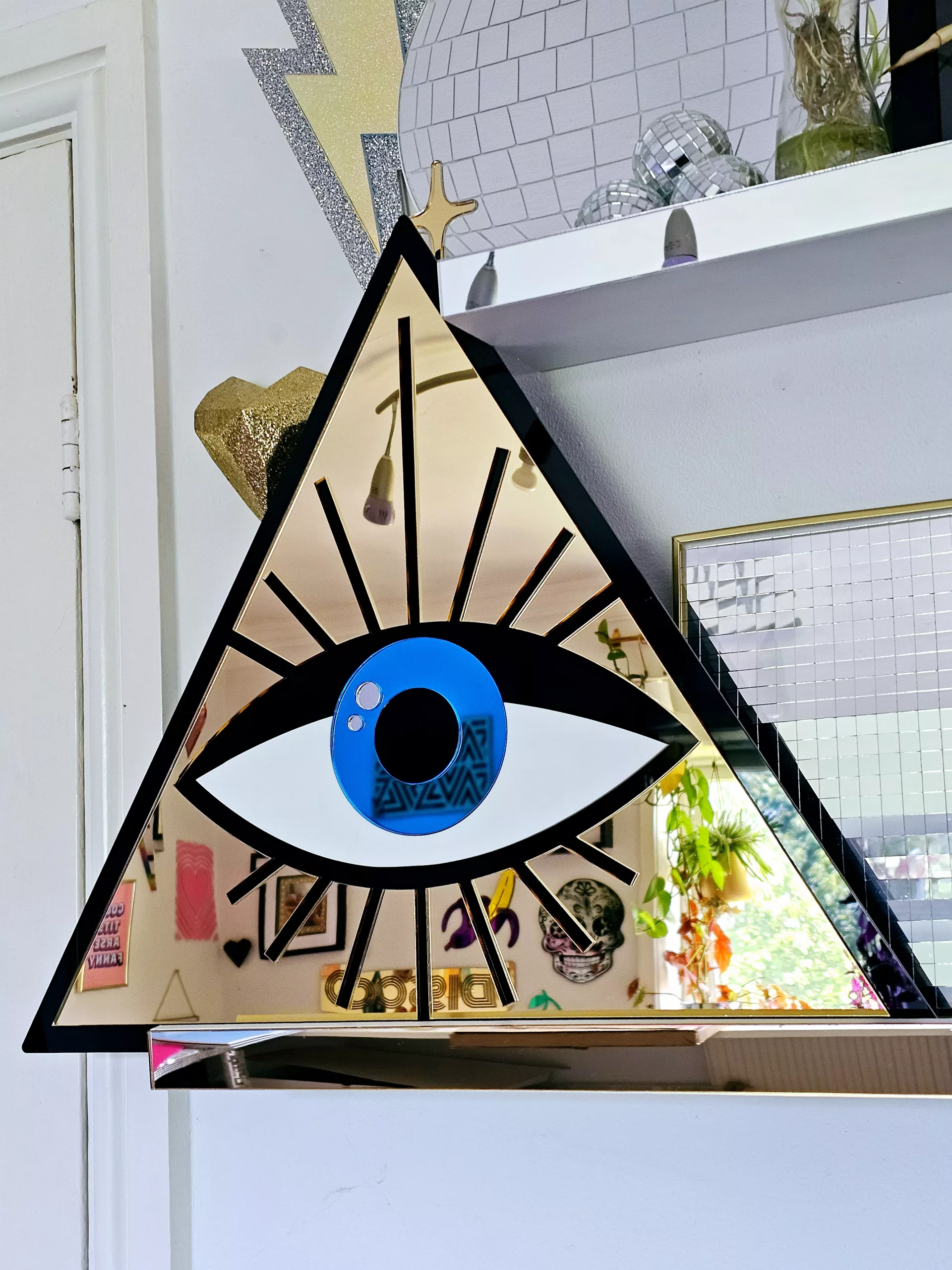 A mirror in the shape of an all seeing eye. The mirror is a gold triangle with an eye in the middle.