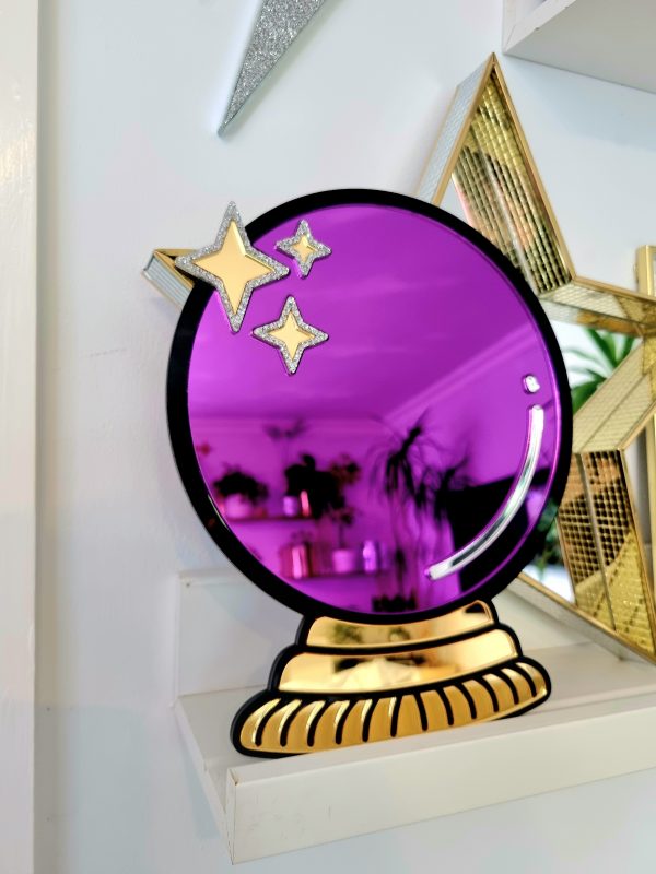 A handmade mirror in the shape of a crystal ball. The design has a black outline with gold mirror stand and purple mirror crystal ball.