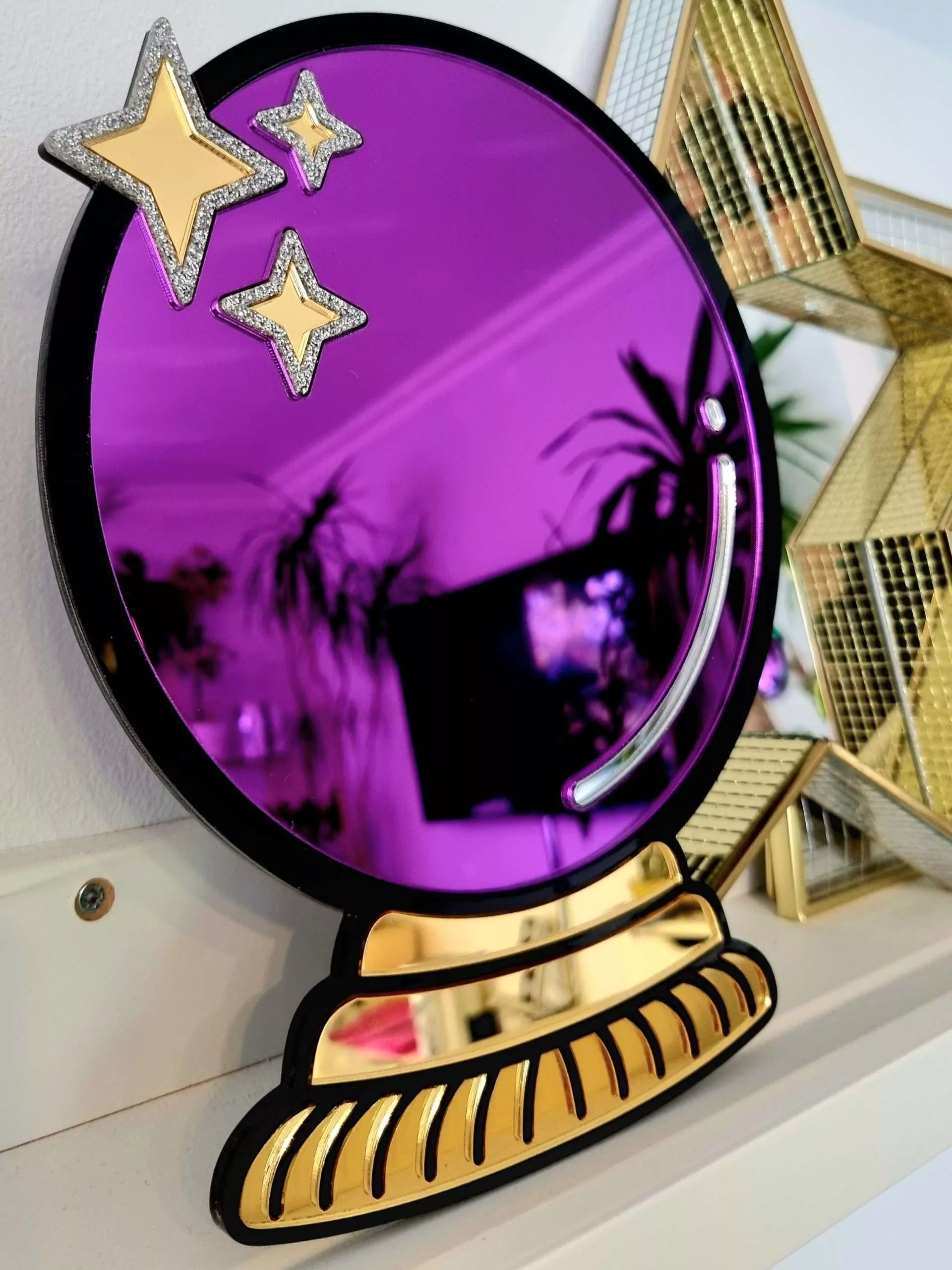 A handmade mirror in the shape of a crystal ball. The design has a black outline with gold mirror stand and purple mirror crystal ball.
