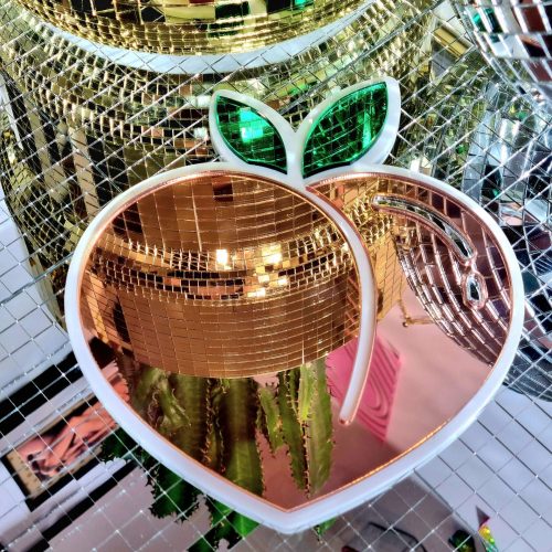 A mirror in the design of the icomic peack emoji. The mirror has a white glossy base and pieces of peach and green mirror.