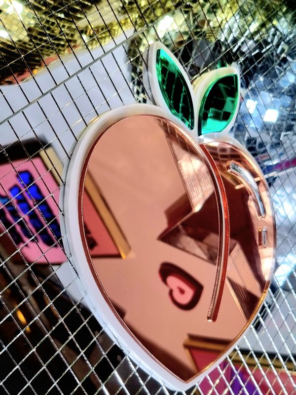 A mirror in the design of the icomic peack emoji. The mirror has a white glossy base and pieces of peach and green mirror.