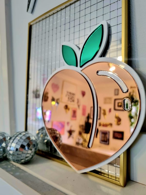 A mirror in the design of the icomic peack emoji. The mirror has a white glossy base and pieces of peach and green mirror.