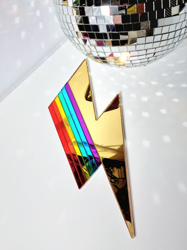 A handmade mirror in the shape of a lightning bolt. Most of the bolt is silver, with strips representing each colour of the rainbow.