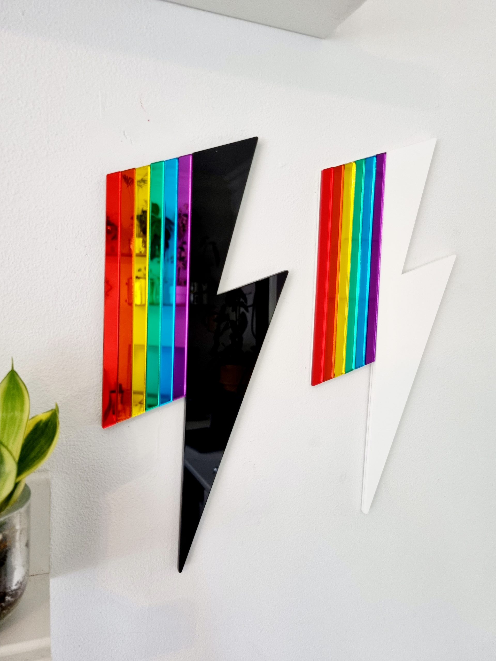 Two pieces of wall art in the shape of lightning bolts. Each has either a white or black gloss base, accented with pieces or rainbow coloured mirror.