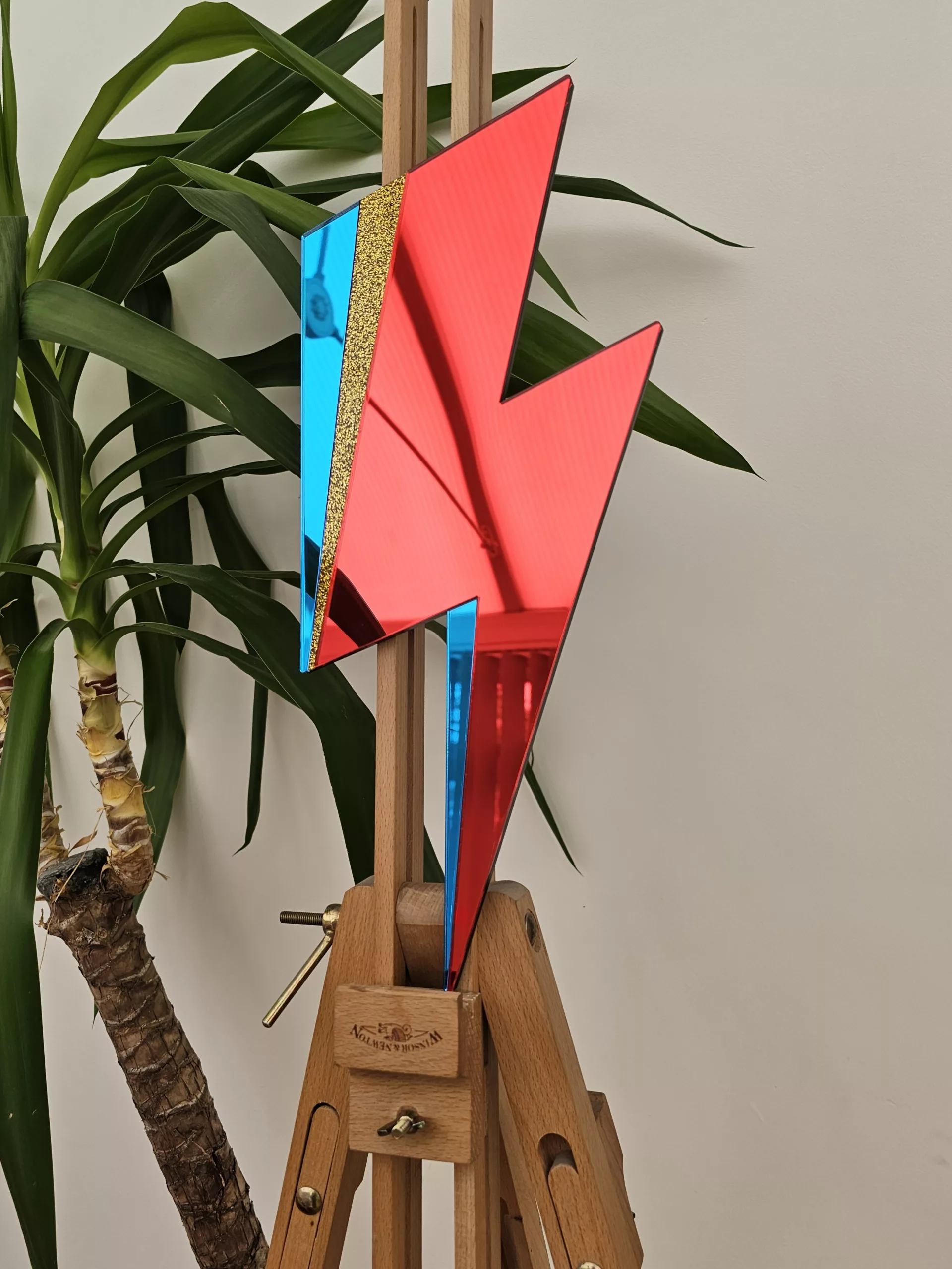 A handmade mirror in the style of the Aladdin Sane David Bowie lghtning bolt. The bolt is made from red and blue mirror with a flash of gold glitter.