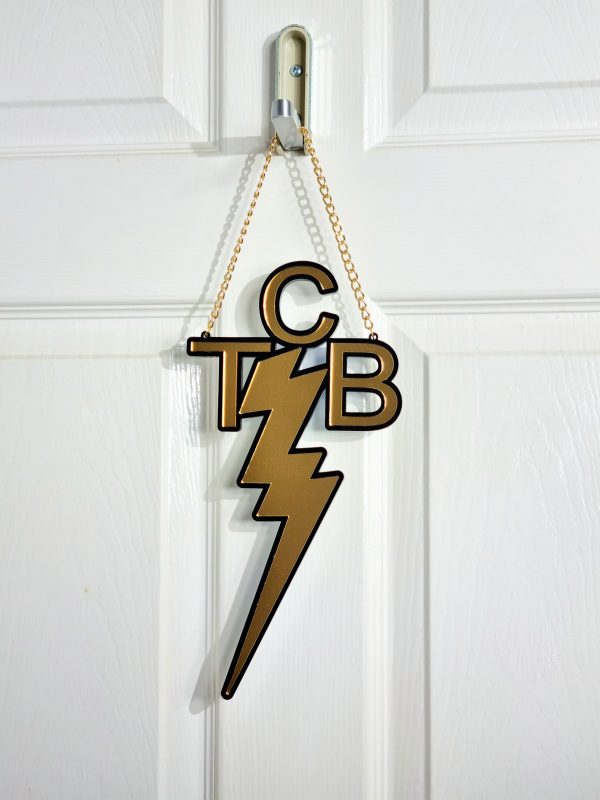 A handmade wall or door hanging in the style ofthe TCB necklace worn by Elvis Presley. The piece is gold with a black outline.
