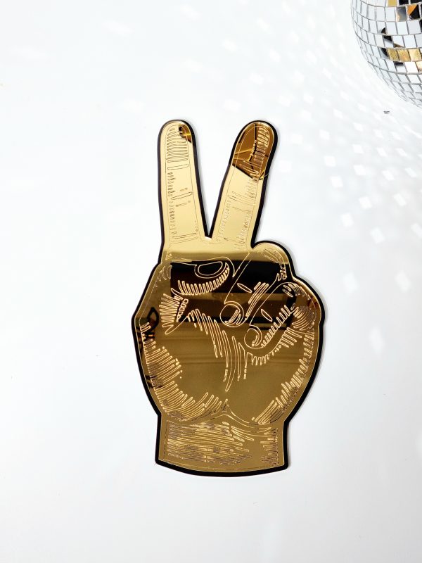 A gold mirror in the shape of a hand giving the peace sign. The mirror is gold with a black outline and has details engraved from underneath.