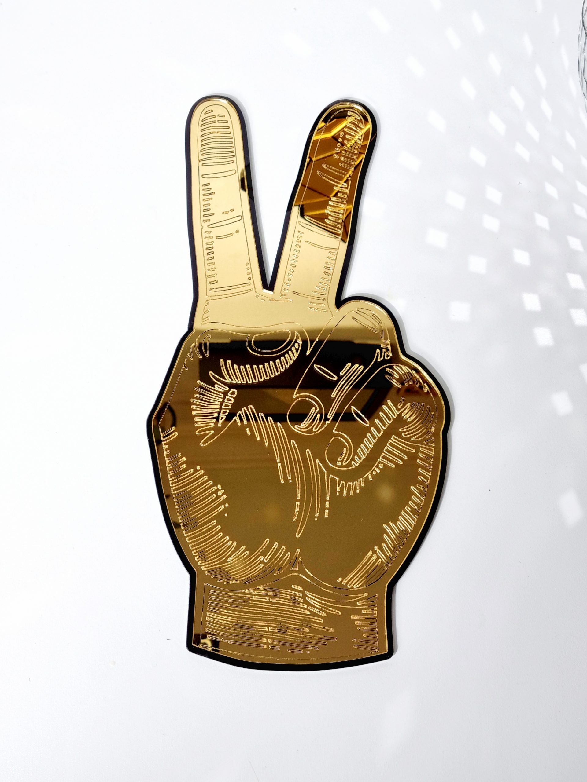 A gold mirror in the shape of a hand giving the peace sign. The mirror is gold with a black outline and has details engraved from underneath.