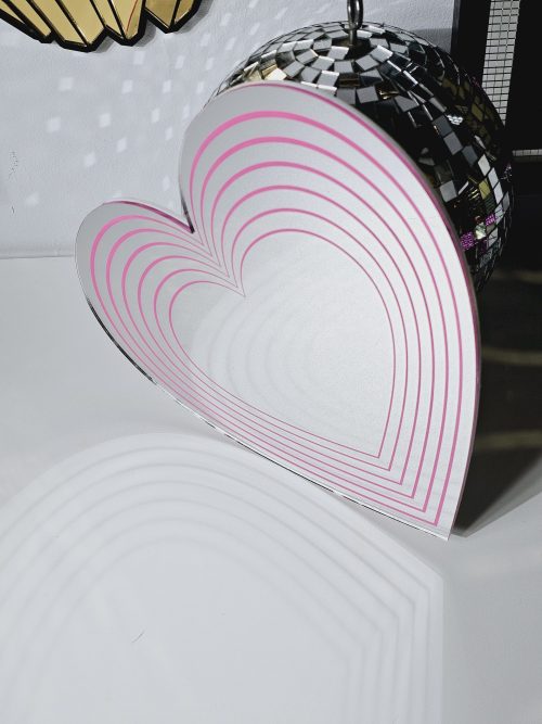 A silver mirror in the shape of a heart with pink hearts of decreasing sizes within it.