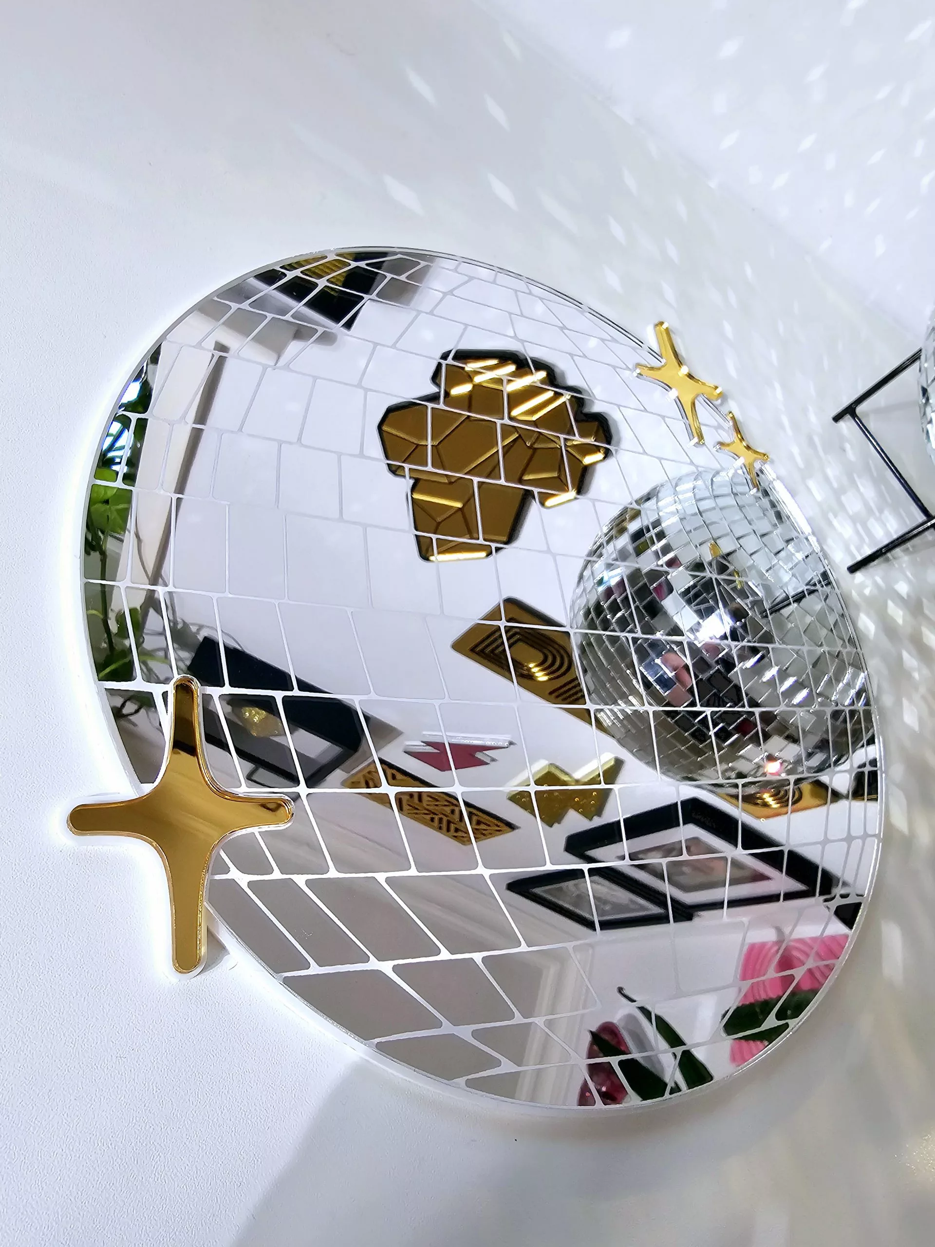 A handmade mirror in the shape of a disco ball. The mirror is silver with white lining and gold twinkles.