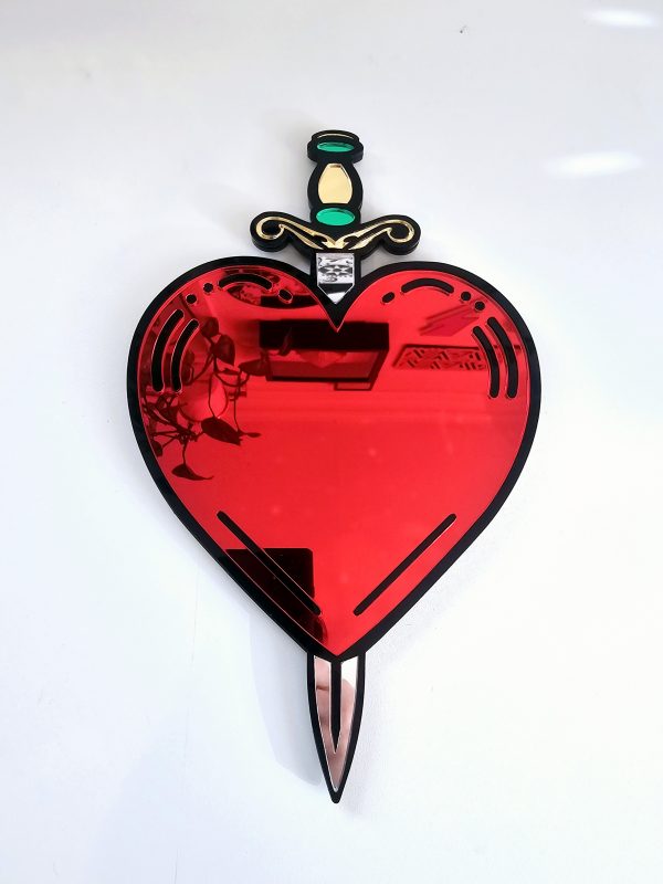 A handmade mirror in the shape of a heart and dagger, in an old school tattoo style. The heart is red with a black outline and orgate dagger design.