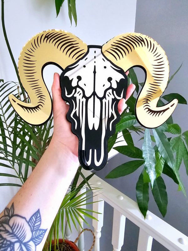 A mirror in the shape of a ram skull. The piece has gold mirrored horns and a silver mirror facee with black detailing.