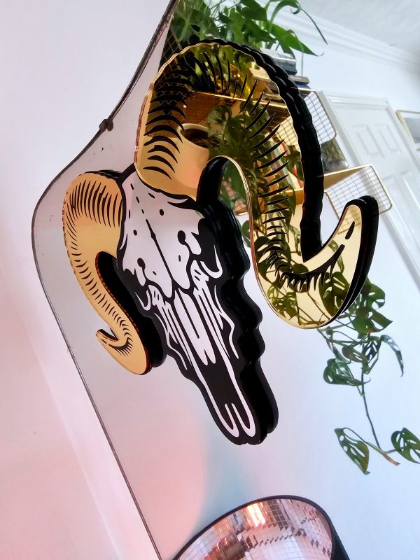 A mirror in the shape of a ram skull. The piece has gold mirrored horns and a silver mirror facee with black detailing.