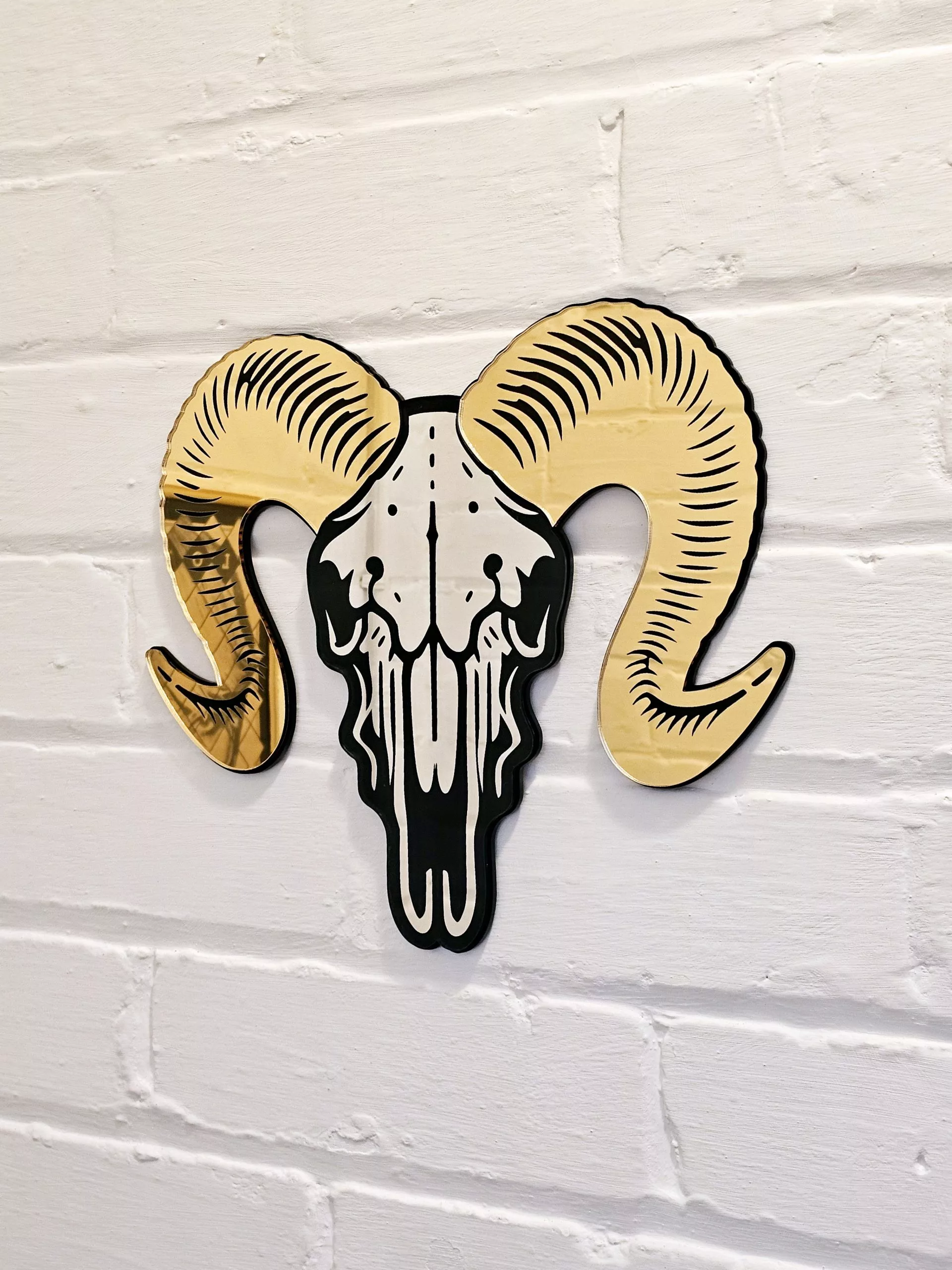 A mirror in the shape of a ram skull. The piece has gold mirrored horns and a silver mirror facee with black detailing.