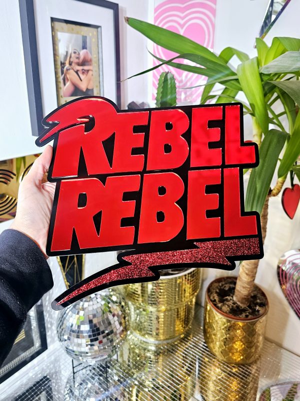 A piece of wall art with the text Rebel Rebel in red mirror with a black background.