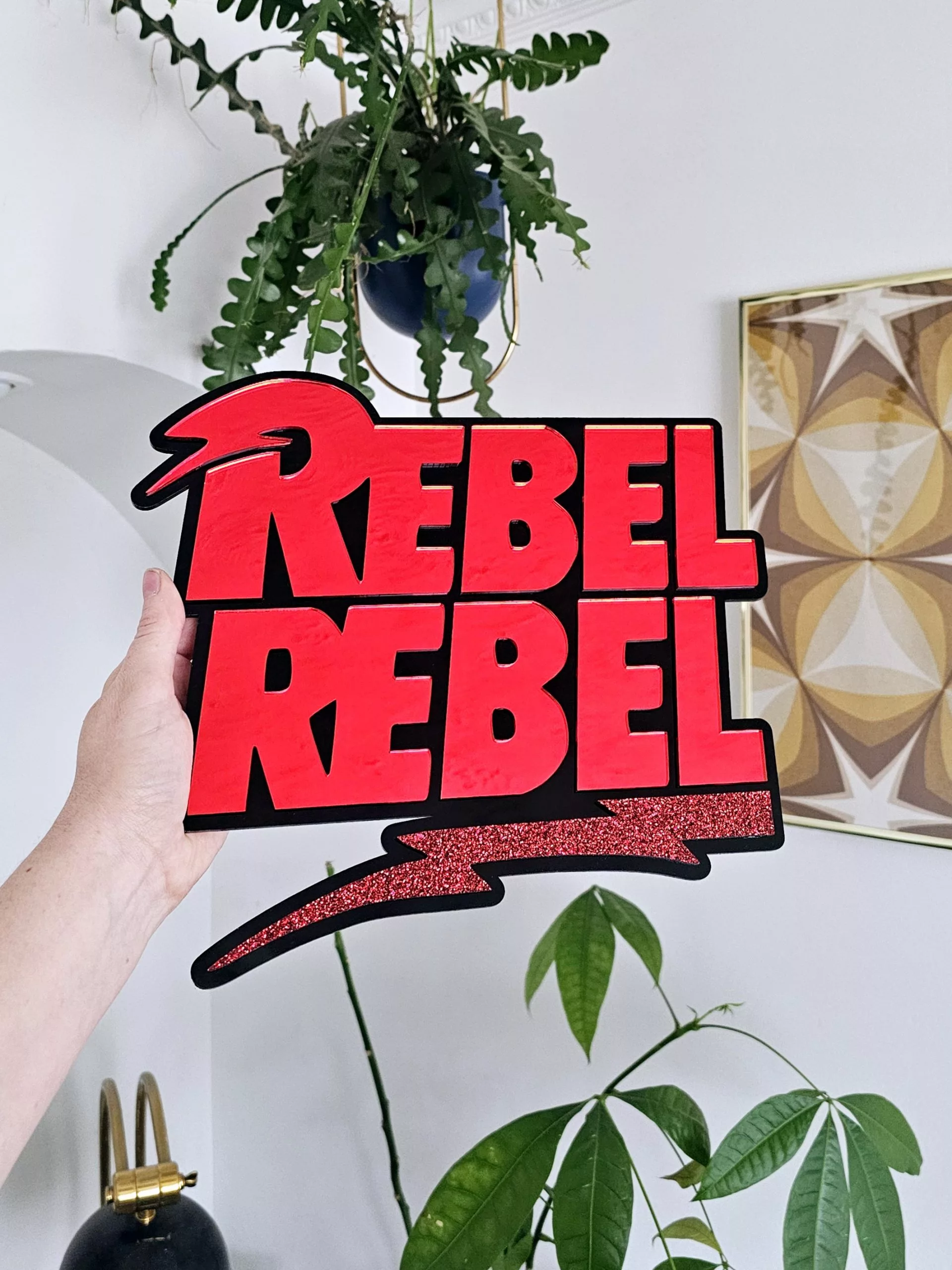 A piece of wall art with the text Rebel Rebel in red mirror with a black background.