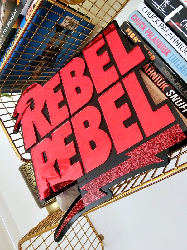 A piece of wall art with the text Rebel Rebel in red mirror with a black background.