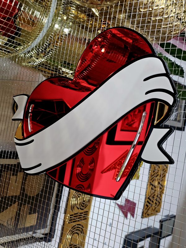 A handmade mirror in the style of a traditional tattoo heart with banner through the middle. The heartis red with a black outline and white banner with silver and gold mirror accents.