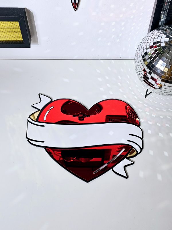 A handmade mirror in the style of a traditional tattoo heart with banner through the middle. The heartis red with a black outline and white banner with silver and gold mirror accents.