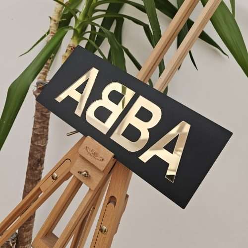 A piece of handmade wall art made with matt black acrylic and the word ABBA in gold mirror lettering.