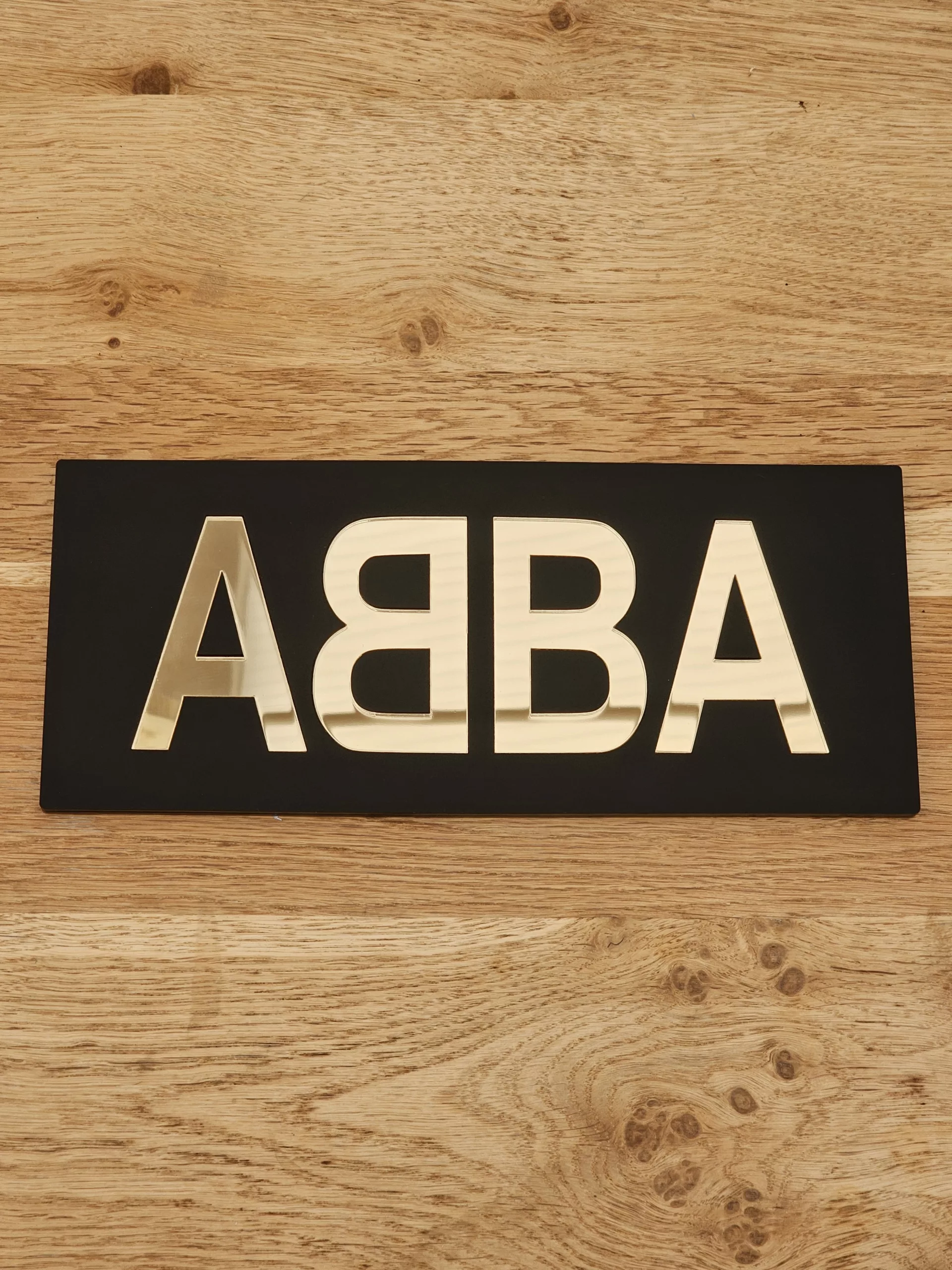 A piece of handmade wall art made with matt black acrylic and the word ABBA in gold mirror lettering.