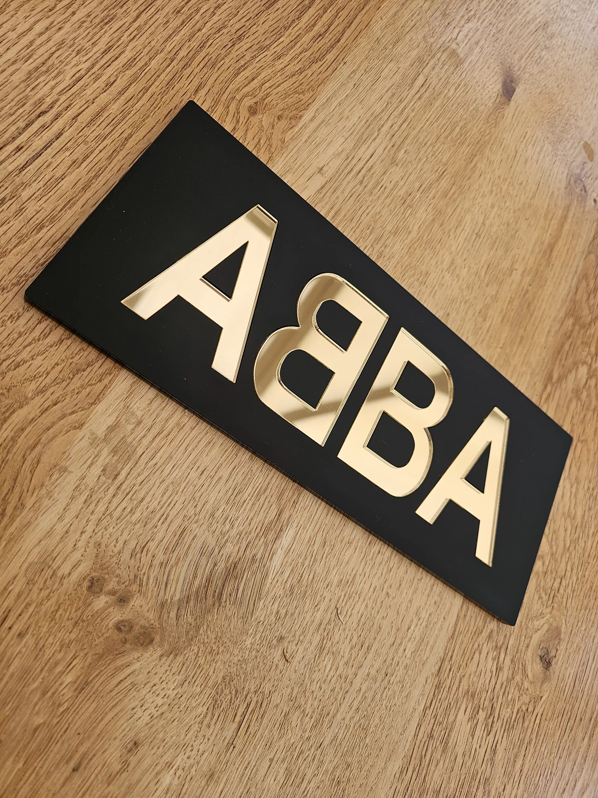 A piece of handmade wall art made with matt black acrylic and the word ABBA in gold mirror lettering.