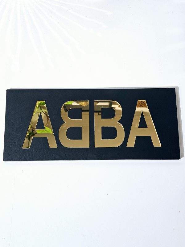A piece of handmade wall art made with matt black acrylic and the word ABBA in gold mirror lettering.