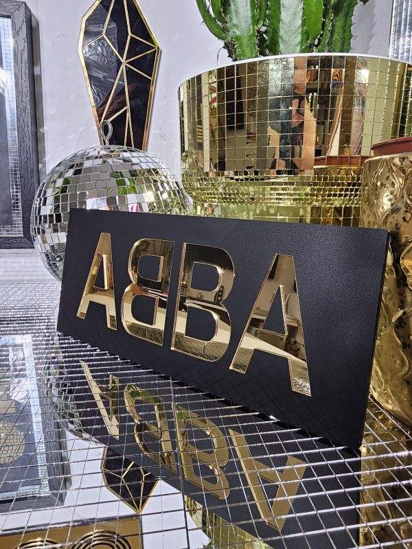 A piece of handmade wall art made with matt black acrylic and the word ABBA in gold mirror lettering.