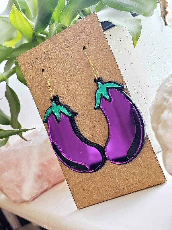 A pair of earrings in the shape of an aubergine. The earrings are purple with a black outline and green details.
