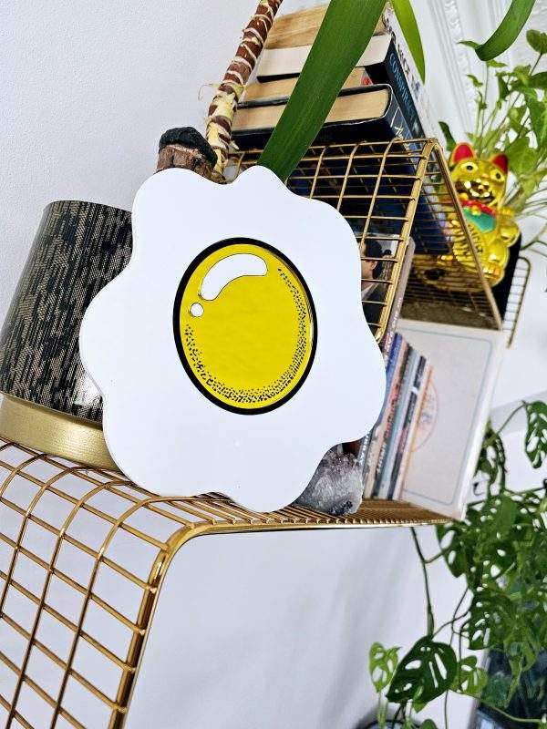 A piece of hand made wall art in the shape of an egg with a yellow mirrored yolk. The piece is sitting on wgold wire shelf.