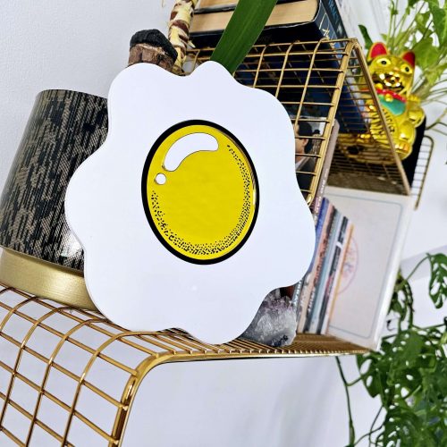 A piece of hand made wall art in the shape of an egg with a yellow mirrored yolk. The piece is sitting on wgold wire shelf.