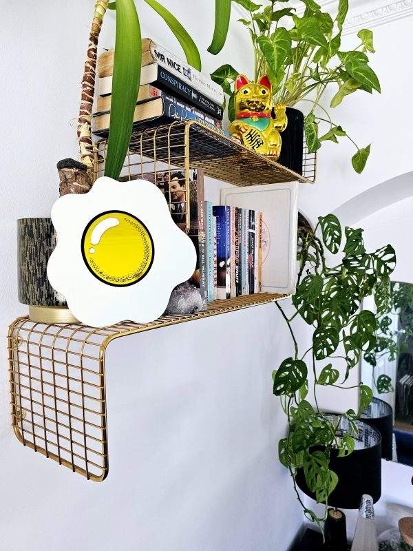 A piece of hand made wall art in the shape of an egg with a yellow mirrored yolk. The piece is sitting on wgold wire shelf.