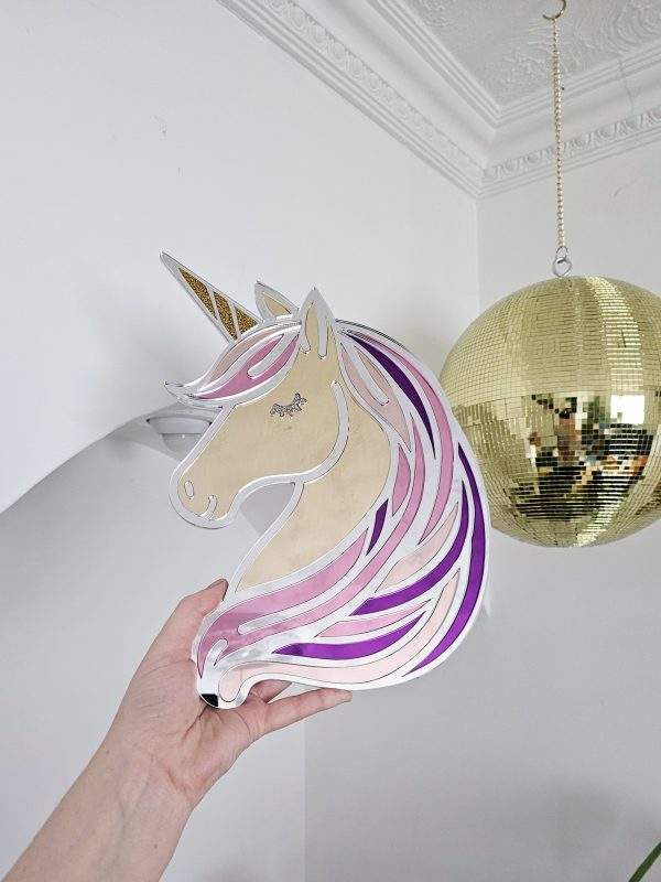 A mirror in the shape of a unicorn head. The unicorn has a silver mane with flecks of pink, purple and rose gold, with glitter horn and eyes.