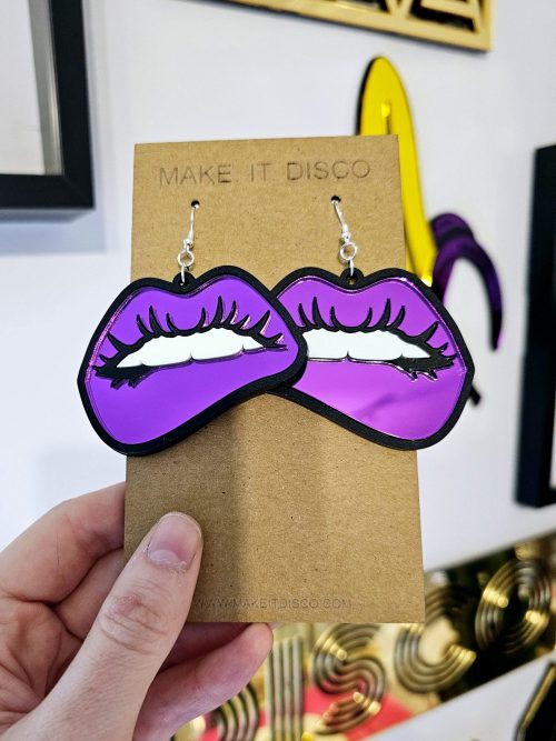A pair of earrings in the shape of lips. They're in a pop art style with black outline and purple mirror lips.