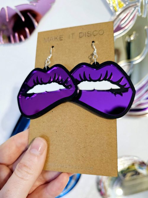 A pair of earrings in the shape of lips. They're in a pop art style with black outline and purple mirror lips.
