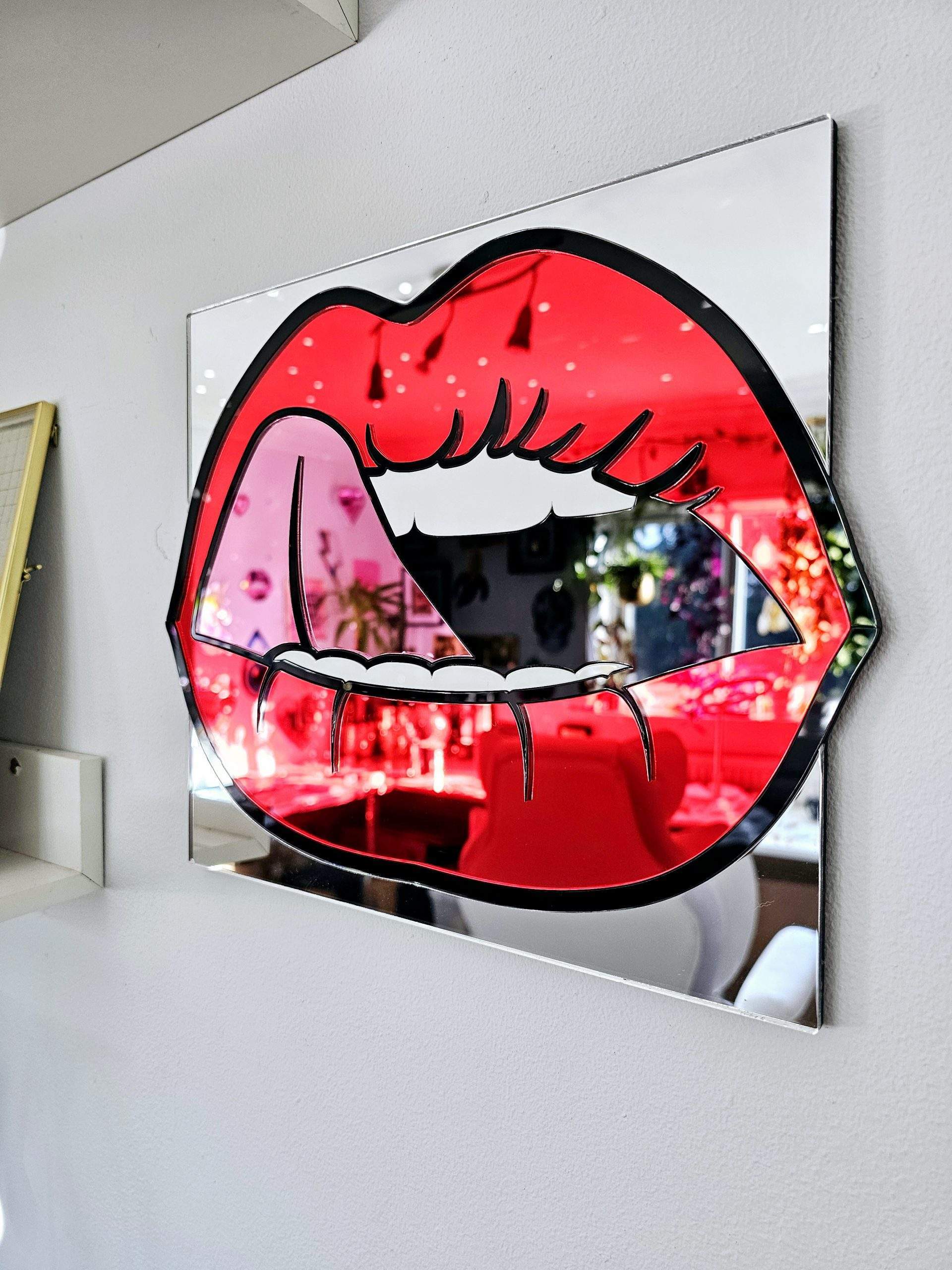 A mirror in the shape of lips. The lips are made in a pop art style with red mirror lips, a pink tongue and black outline.