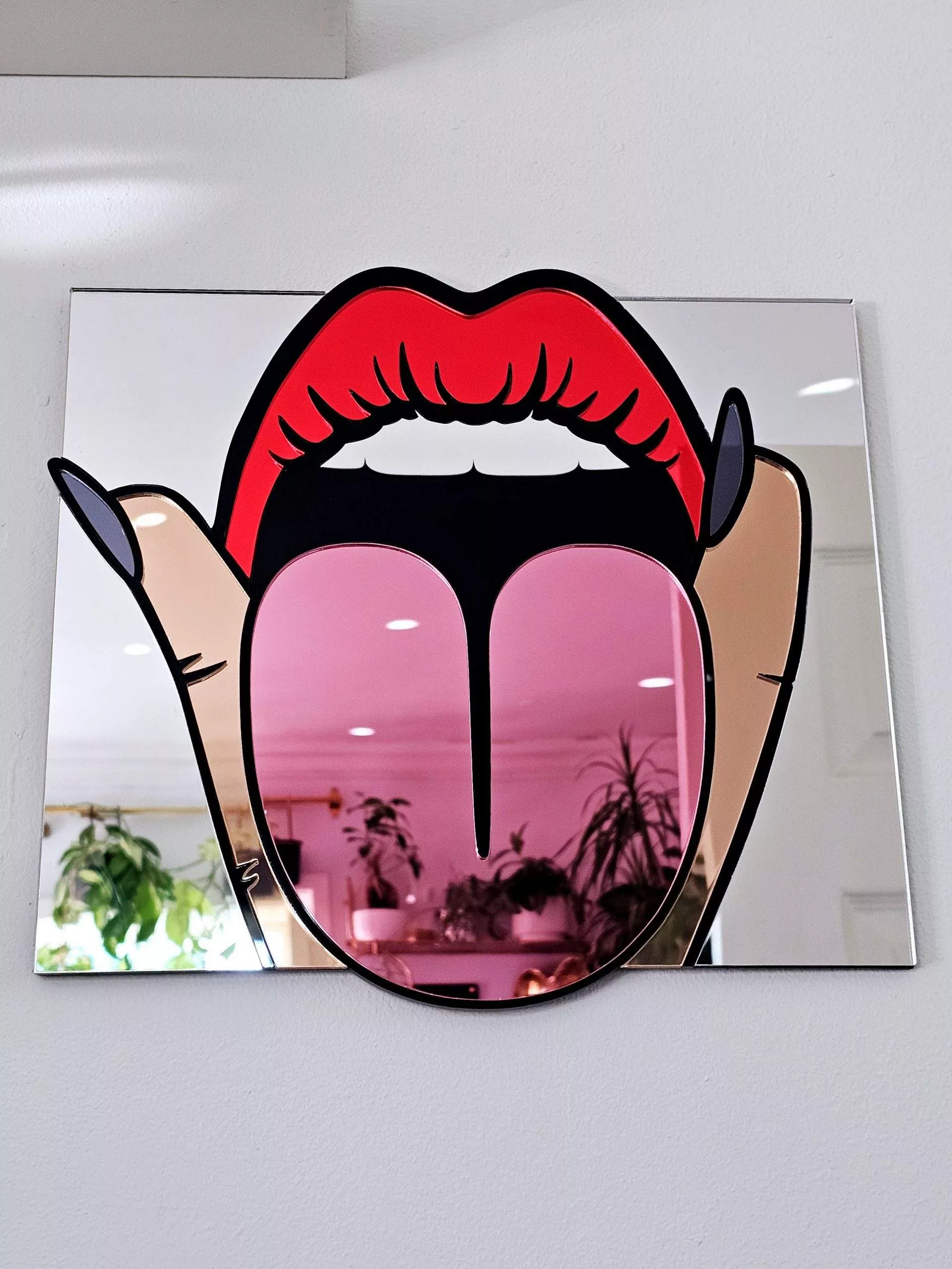A handmade mirror in the shape of a mouth making a rude gesture, with tongue out and fingers either side.