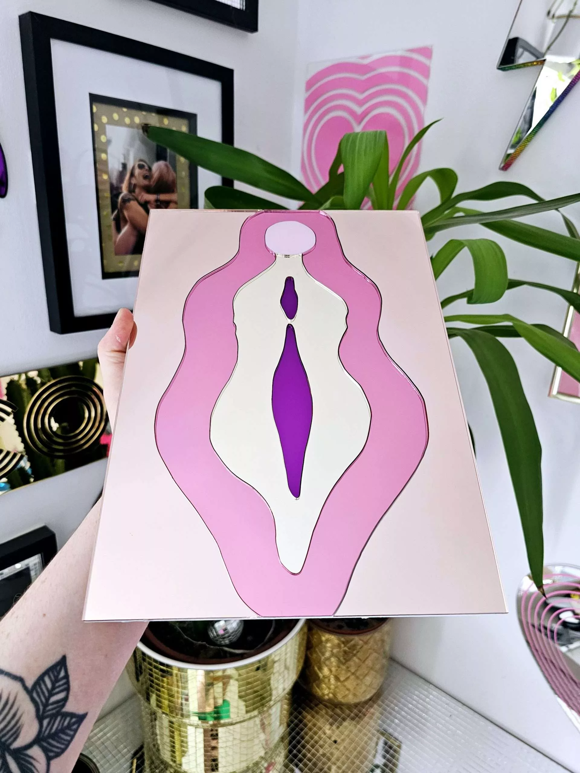 A mirror in the design of a vulva. The mirror is made from pieces of rose gold, pink, gold and purple mirror.