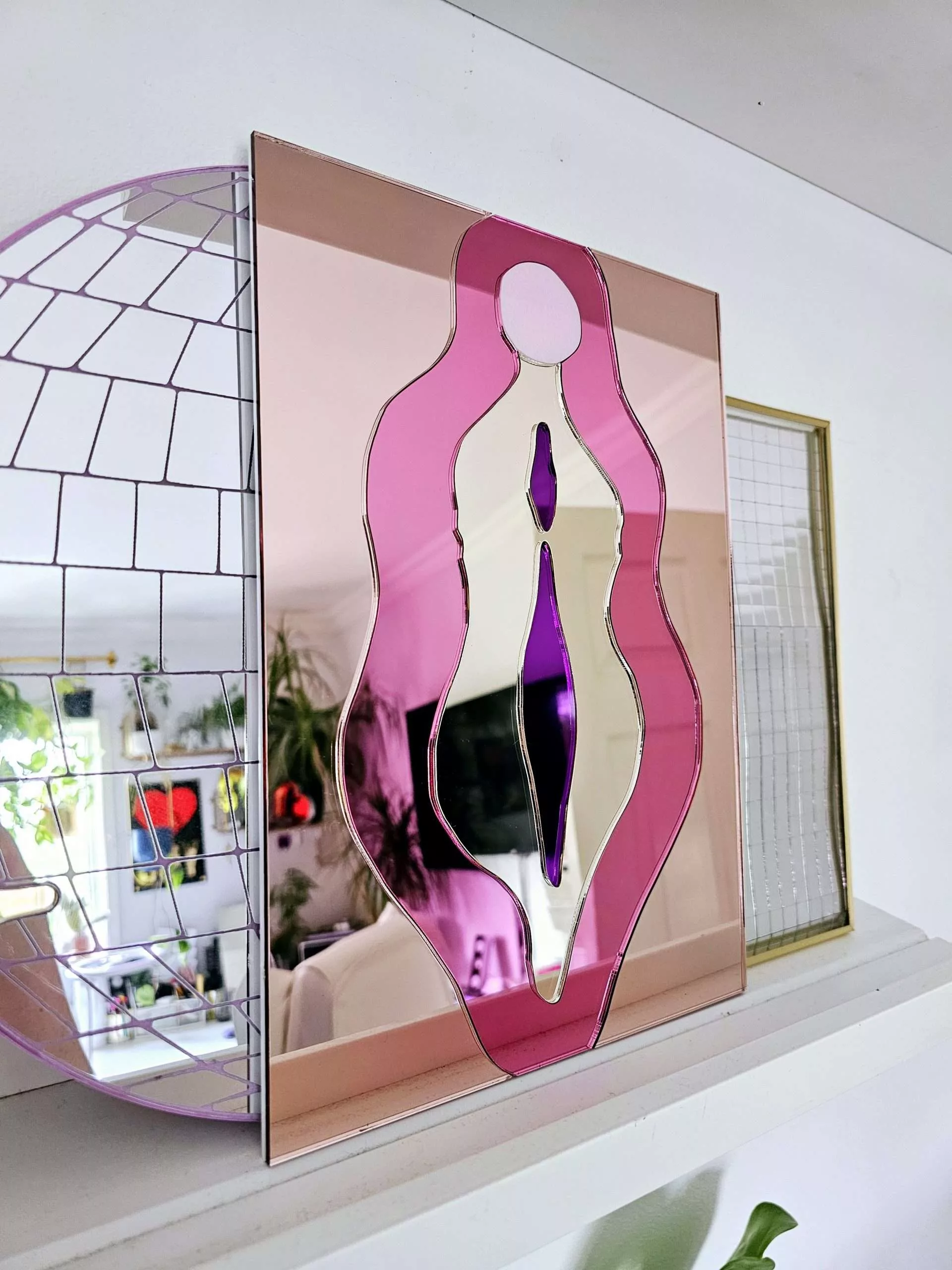 A mirror in the design of a vulva. The mirror is made from pieces of rose gold, pink, gold and purple mirror.