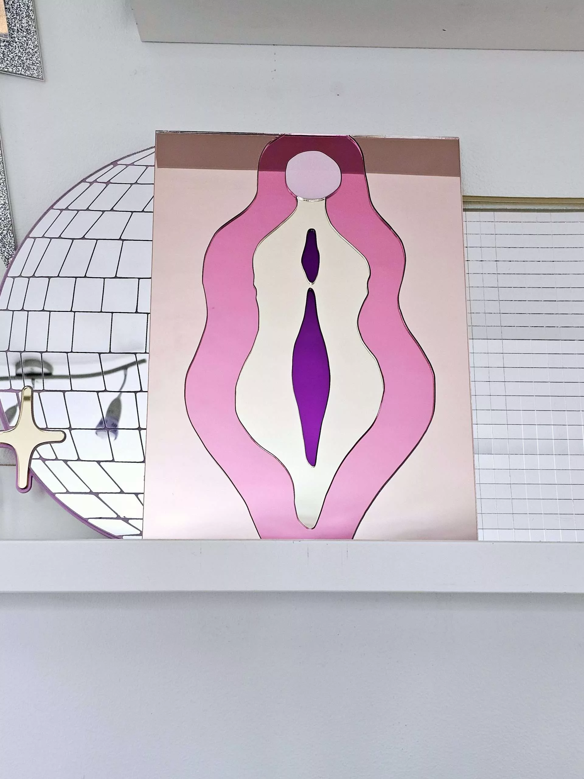 A mirror in the design of a vulva. The mirror is made from pieces of rose gold, pink, gold and purple mirror.