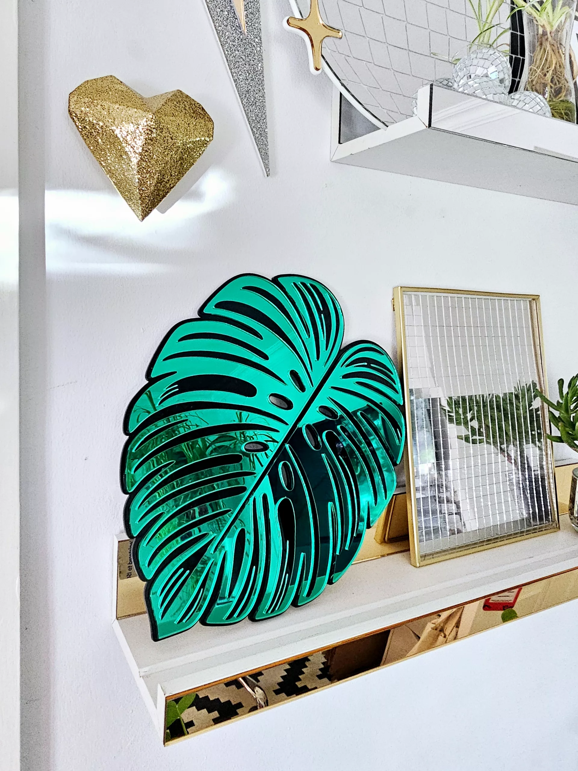 A handmade mirror in the shape of a monstera leaf. The mirror has a black outline, with green and silver mirror leaves.