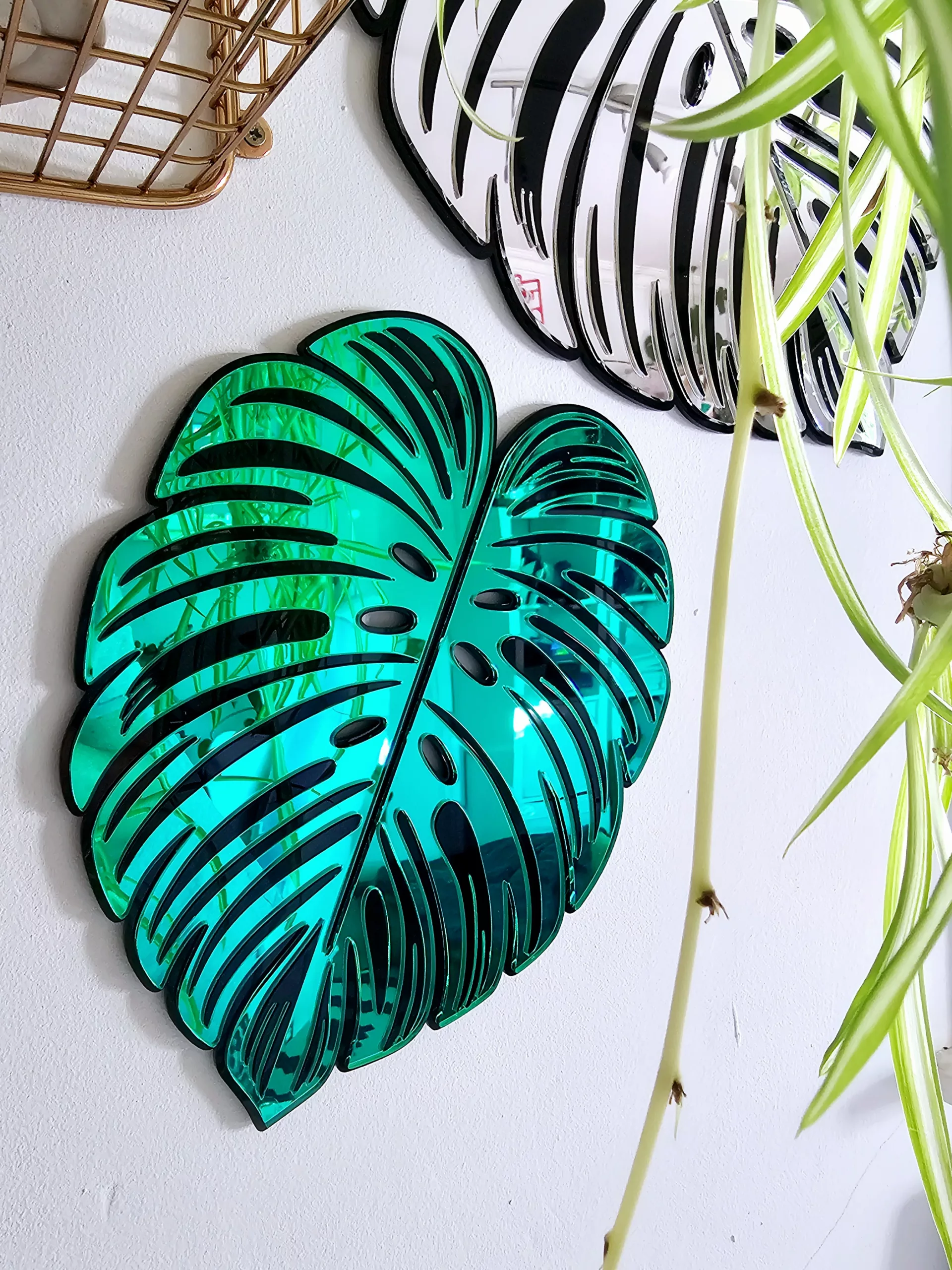 A handmade mirror in the shape of a monstera leaf. The mirror has a black outline, with green and silver mirror leaves.