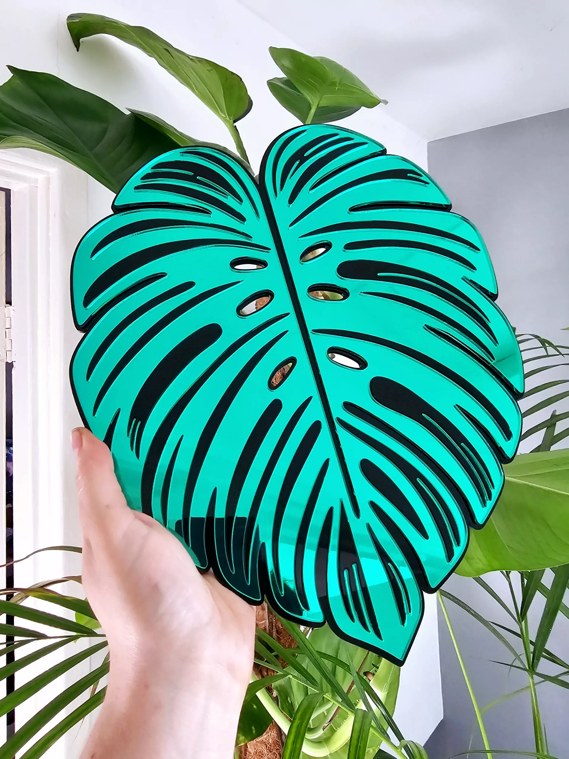 A handmade mirror in the shape of a monstera leaf. The mirror has a black outline, with green and silver mirror leaves.