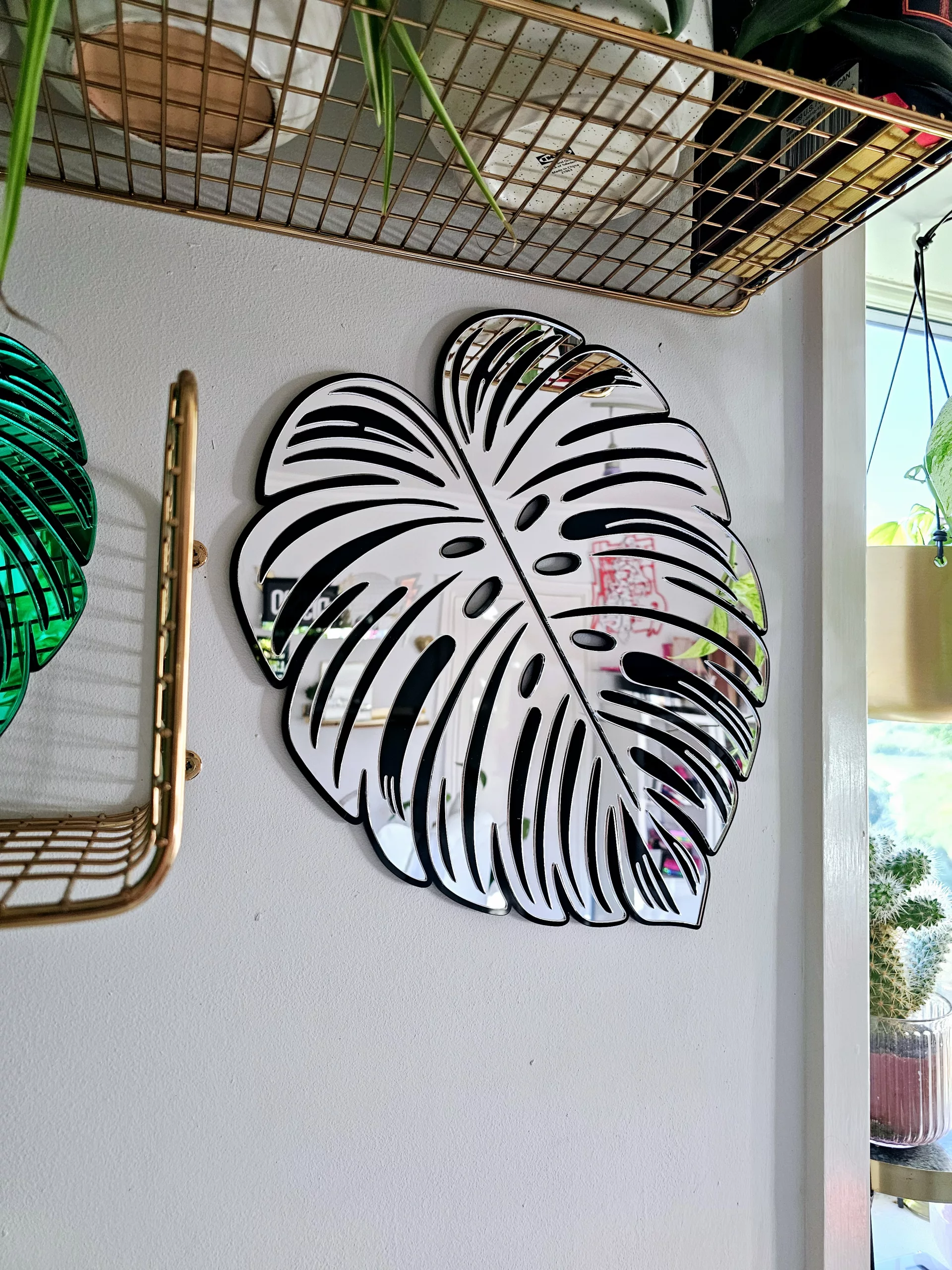A handmade mirror in the shape of a monstera leaf. The mirror has a black outline, with green and silver mirror leaves.