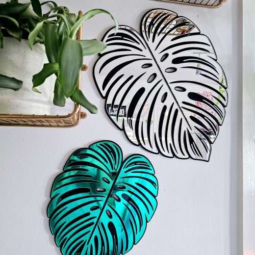 A handmade mirror in the shape of a monstera leaf. The mirror has a black outline, with green and silver mirror leaves.
