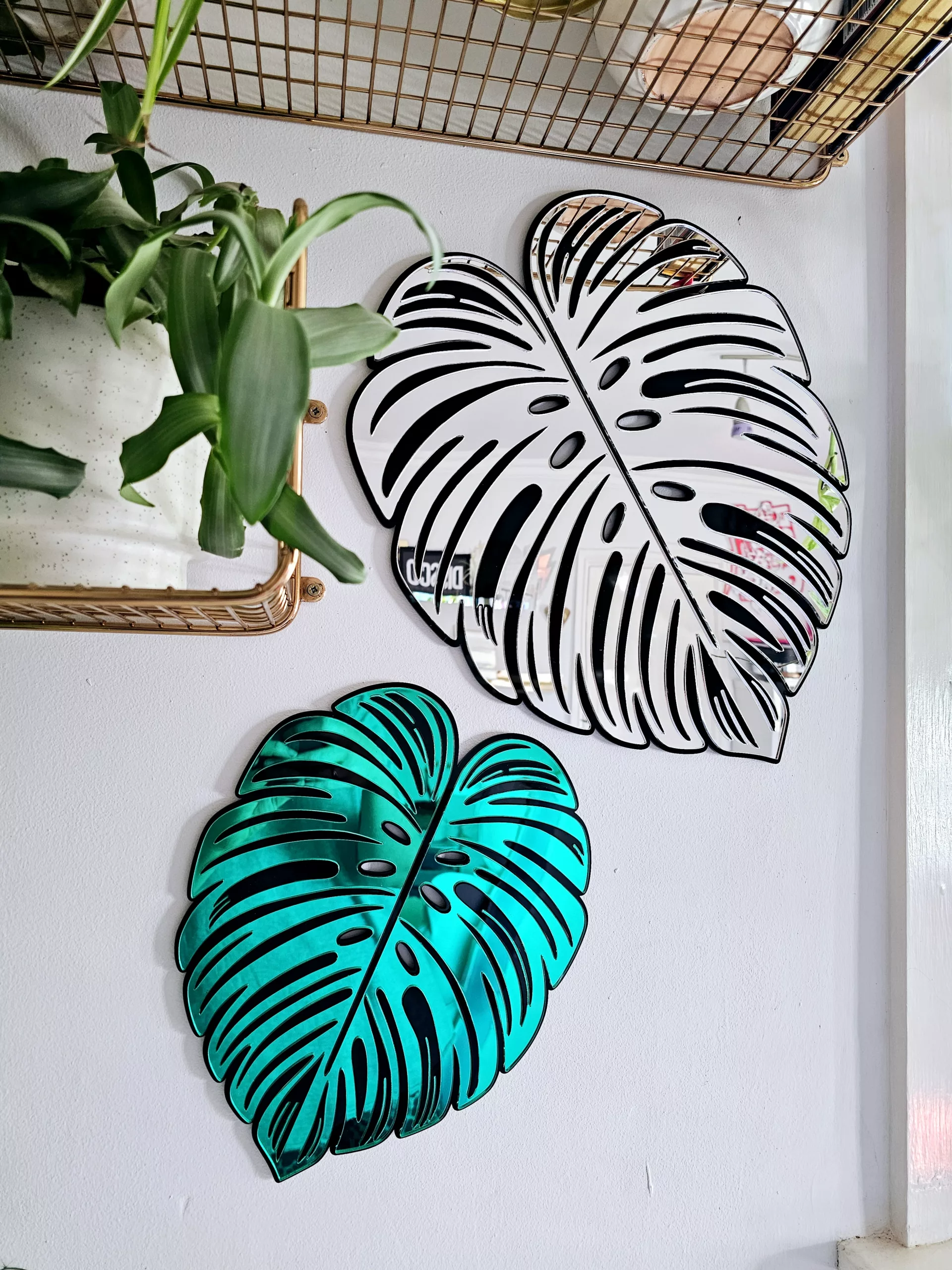 A handmade mirror in the shape of a monstera leaf. The mirror has a black outline, with green and silver mirror leaves.
