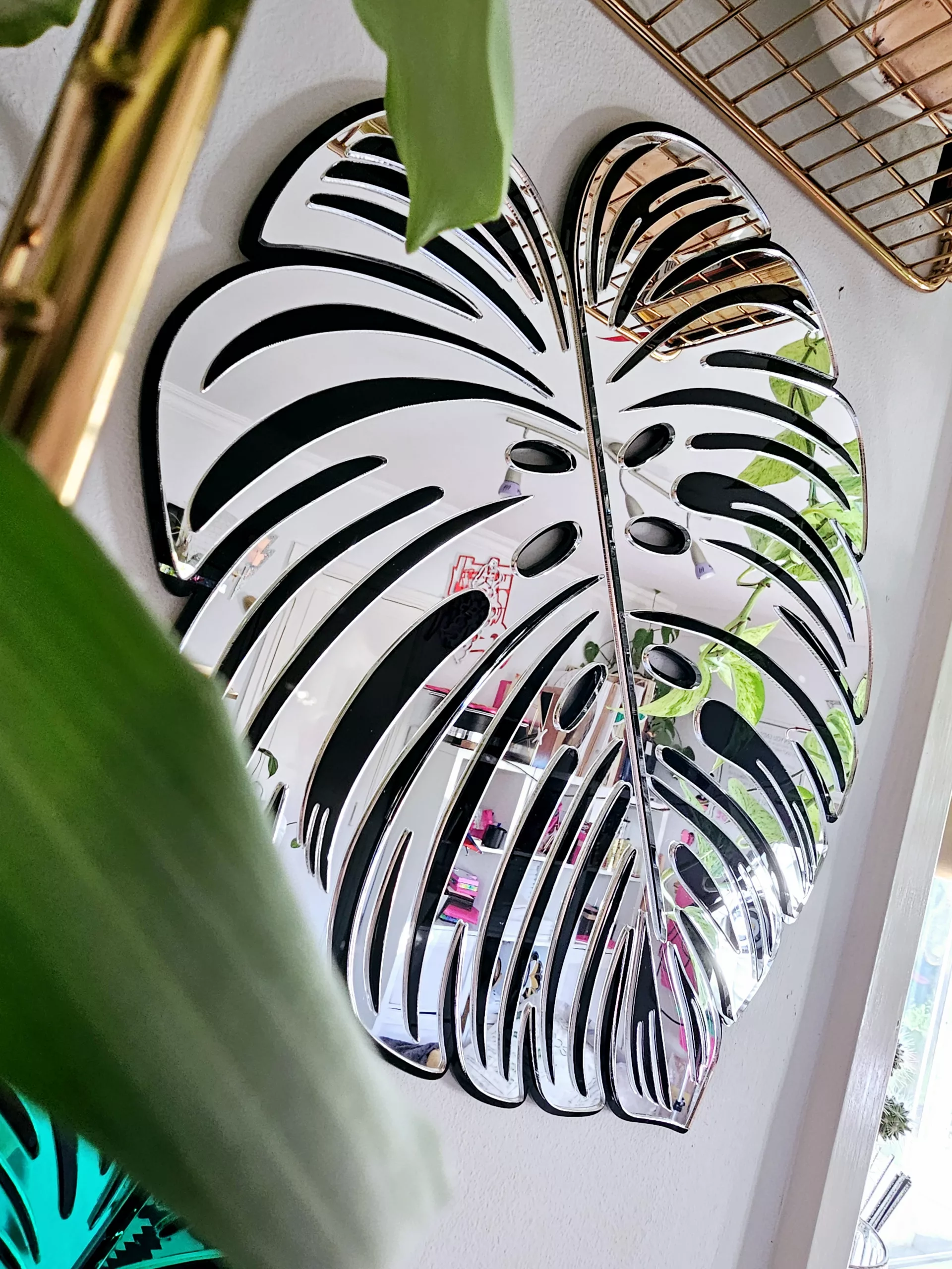 A handmade mirror in the shape of a monstera leaf. The mirror has a black outline, with silver mirror leaves.