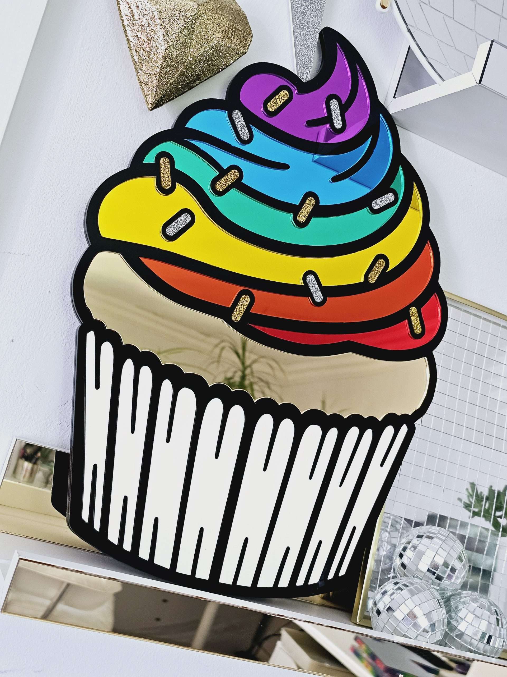 A piece of wall art in the shape of a cupcake. The cupcake has a black outline, rainbow mirror frosting with glitter sprinkles and a matt white cupcake case.