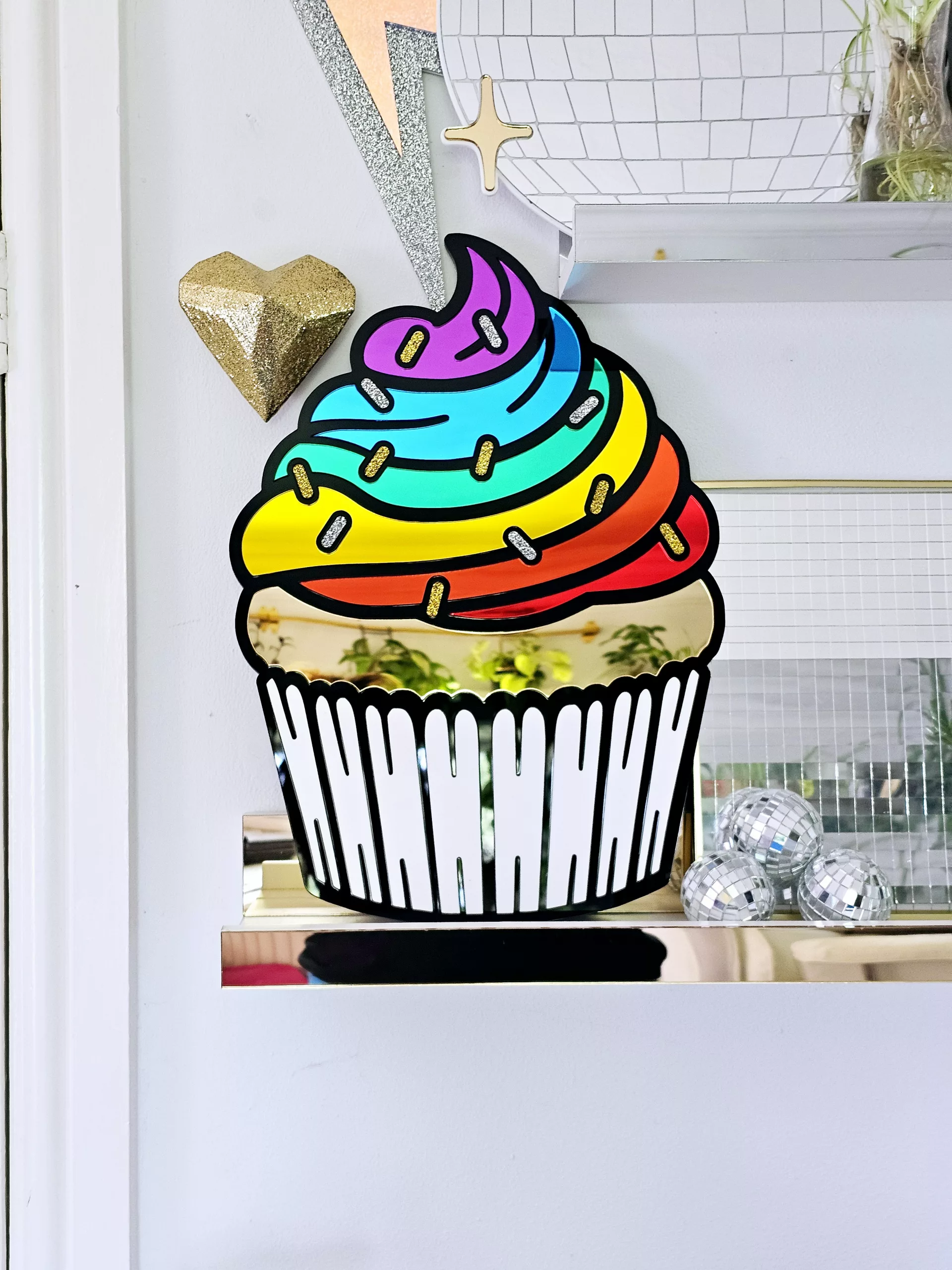 A piece of wall art in the shape of a cupcake. The cupcake has a black outline, rainbow mirror frosting with glitter sprinkles and a matt white cupcake case.
